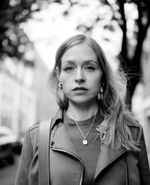 Deri ceket ve kolye giyen, arka planda bulanık ağaçlar ve binalar olan şehir ortamında poz veren sarışın genç bir kadının tek renkli orta formatlı film portresi.