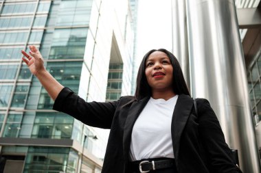 İş kadını, hareketli bir şehir iş bölgesinde taksiyi selamlıyor, etkileyici gökdelenler ve modern mimari ile çevriliyken güvenli bir şekilde ulaşımı işaret ediyor.