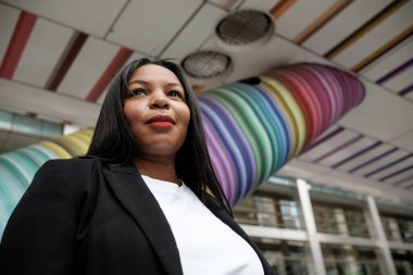 İş kadını canlı, gökkuşağı renginde bir mimari tasarımın altında durarak profesyonellik ve modern estetiğin bir karışımını sergileyerek kendine güvenini sergiliyor.