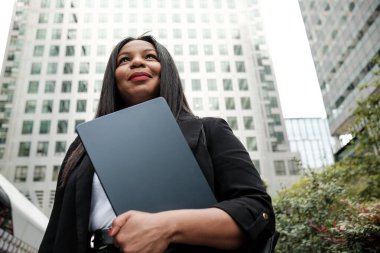 Dizüstü bilgisayar tutan kendine güvenen iş kadını, modern gökdelenlere olumlu bir ifadeyle bakıyor ve iş dünyasında başarıyı öngörüyor.