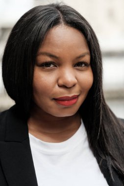 Şık bir spor ceket ve beyaz tişört giyen gülümseyen bir iş kadınının portresi canlı bir şehir planına karşı kendine güven ve profesyonellik yayıyor.