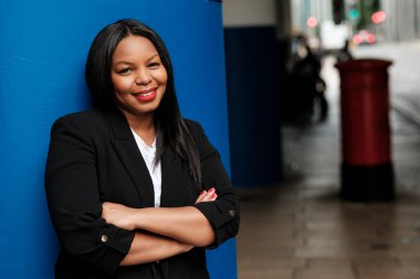 İş kadını, şehir ortamında canlı mavi bir duvara yaslanmış, kollarını kavuşturmuş, yüzünde bir gülümseme ile profesyonellik ve başarının bir görüntüsünü yansıtıyor.