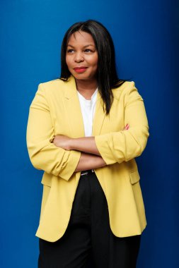 Sarı ceketli ve siyah pantolonlu bir iş kadını, kollarını kavuşturmuş özgüvenini ve yüzünde düşünceli bir gülümsemeyle canlı bir mavi arkaplan karşısında duruyor.