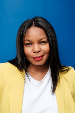 Sarı ceket ve beyaz tişört giyen gülümseyen bir iş kadınının stüdyo portresi, canlı mavi bir arka plana karşı poz veriyor, özgüven ve profesyonellik yayıyor.