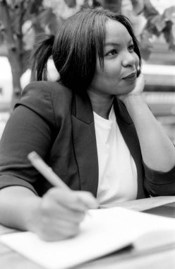 Siyah ve beyaz analog görüntü, bir iş kadınının dış mekan ortamında düşünürken notlar yazması ve sol elinin üzerine başını koyması.