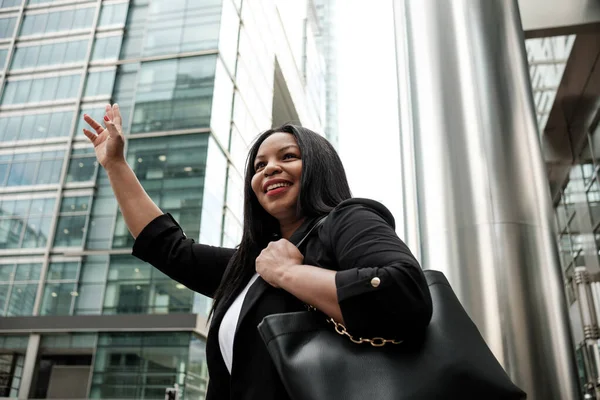 Gülümseyen bir iş kadını şık modern ofis binalarının önünde taksi çağırıyor, şık bir ceket giymiş, kendine güveni ve profesyonelliği olan şık siyah bir çanta tutuyor.
