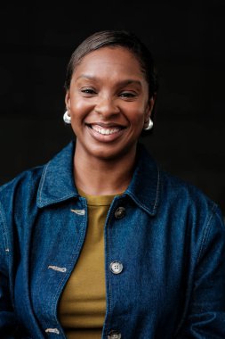 Kot ceket giyen, gülümseyen, özgüven ve pozitiflik saçan, koyu bir arka plana karşı gülümseyen Afrikalı Amerikalı bir kadının stüdyo portresi.