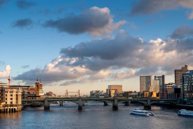 Londra şehir manzarası, nehir boyunca uzanan güney köprüsü ile betimleniyor, uzaktaki ikonik kule köprüsünü ve bulutlu bir gökyüzünün altında yüzen tekneleri gösteriyor.
