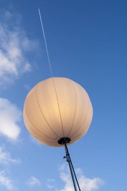 Büyük şeffaf küresel aydınlatma balonu bir tripodun üzerinde duruyor. Parlak mavi bir gökyüzüne karşı. Dağılmış bulutlar ve yukarı doğru uzanan görünür bir uçak izi.