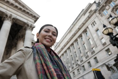 Klasik tarihi binaların altında duran kadın, kameraya gülümsüyor, seyahat ederken atkı ve trençkot giyiyor ve modern şehir mimarisini keşfediyor.