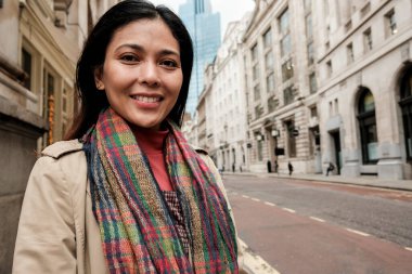 Londra 'da bir caddede kameraya gülümseyen bir kadın. Renkli bir atkı ve yağmurlukla şehir turunun ve güler yüzlü bir sonbahar şehrinin tadını çıkarıyor.