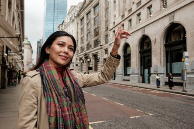 Trençkotlu Asyalı bir kadın Londra caddesinde duruyor. Siyah bir taksiye binmek için elini kaldırırken gülümsüyor. Şehir turunun ve bağımsız seyahatin keyfini çıkarıyor.