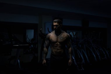 Dövmeli çıplak vücutlu kaslı erkek sporcu. Koyu renk spor salonunda duruyor ve spor yaparken halterli kıvrımlar yapıyor.
