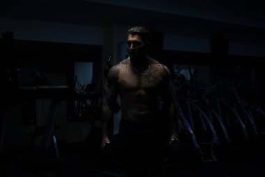 Dövmeli çıplak vücutlu kaslı erkek sporcu. Koyu renk spor salonunda duruyor ve spor yaparken halterli kıvrımlar yapıyor.