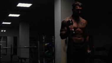 Dövmeli çıplak vücutlu kaslı erkek sporcu. Koyu renk spor salonunda duruyor ve spor yaparken halterli kıvrımlar yapıyor.