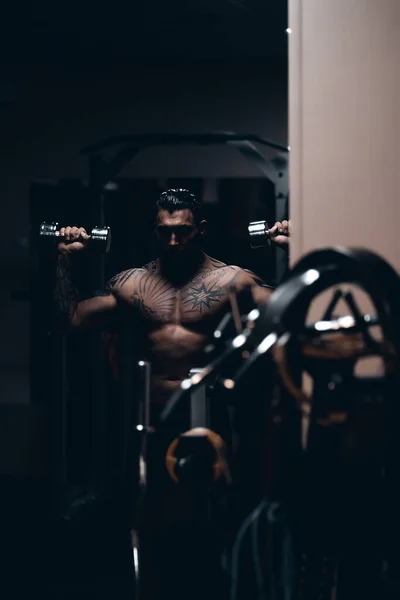 肌肉发达的男性运动员 身体上有纹身 赤身裸体地站在黑暗的体育馆里 在运动时做哑铃卷曲 — 图库照片