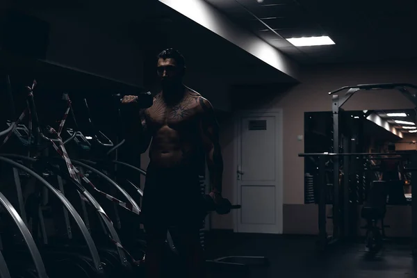 肌肉发达的男性运动员 身体上有纹身 赤身裸体地站在黑暗的体育馆里 在运动时做哑铃卷曲 — 图库照片