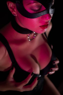 Maskeli ve yakalı isimsiz bir kadın karanlık odadaki BDSM seansında göğüslerine dokunuyor.