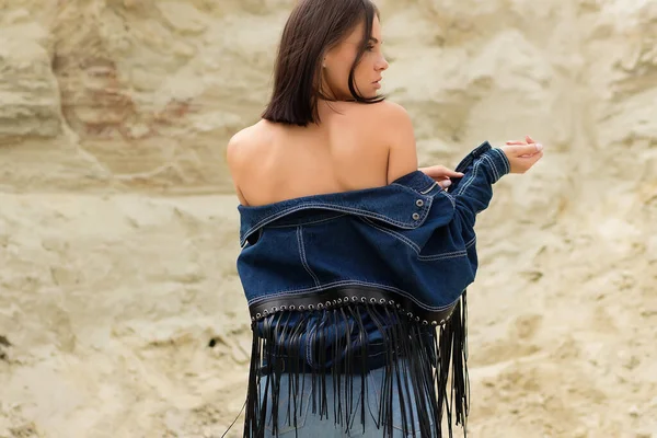 Attractive female model in jeans looking at camera and covering breast with denim jacket on blurred background of countryside