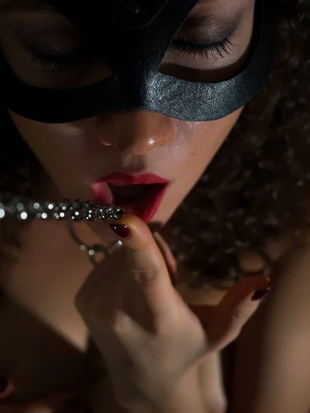 Seductive topless brunette with red lips wearing black mask and metal chain on neck playing BDSM game in darkness