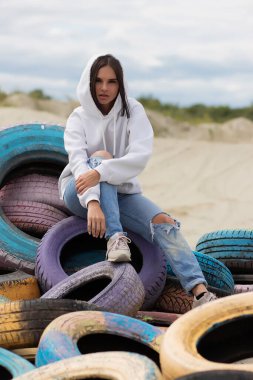 Moda kıyafetli genç bir kadın rüzgarlı bir günde kumsalda dinlenirken kameraya bakıyor ve kapüşonlunun altına dokunuyor.