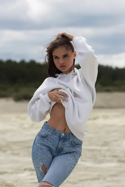 Young woman in trendy outfit looking at camera and touching belly under hoodie while resting on beach on windy day