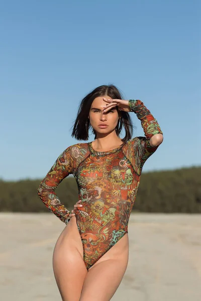 若いですスリム女性で装飾ボディスーツは髪に触れながら 砂のビーチで晴れた日に休みます — ストック写真