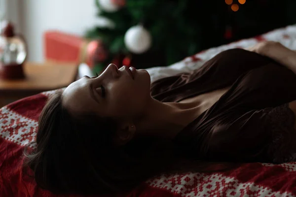 サイドビューの性的若いです女性とともに長い黒髪でエレガントな赤のブラジャー横になって快適なベッドで装飾されたクリスマスツリーの近くに目 — ストック写真