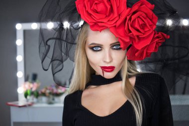 Dudaklarında kan lekesi olan çekici bir kadın Gotik kıyafetler giyip stüdyoda dikilirken kameraya bakıp sırıtıyor.