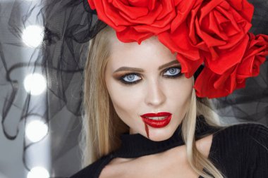 Dudaklarında kan lekesi olan çekici bir kadın Gotik kıyafetler giyip stüdyoda dikilirken kameraya bakıp sırıtıyor.