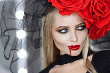 Dudaklarında kan lekesi olan çekici bir kadın Gotik kıyafetler giyip stüdyoda dikilirken kameraya bakıp sırıtıyor.