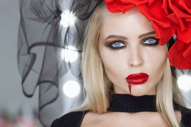 Dudaklarında kan lekesi olan çekici bir kadın Gotik kıyafetler giyip stüdyoda dikilirken kameraya bakıp sırıtıyor.