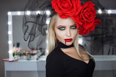 Dudaklarında kan lekesi olan çekici bir kadın Gotik kıyafetler giyip stüdyoda dikilirken kameraya bakıp sırıtıyor.