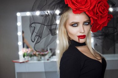 Dudaklarında kan lekesi olan çekici bir kadın Gotik kıyafetler giyip stüdyoda dikilirken kameraya bakıp sırıtıyor.