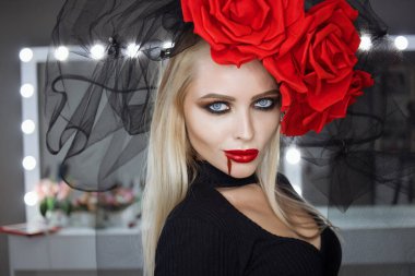 Dudaklarında kan lekesi olan çekici bir kadın Gotik kıyafetler giyip stüdyoda dikilirken kameraya bakıp sırıtıyor.