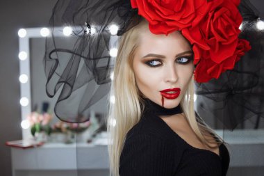 Dudaklarında kan lekesi olan çekici bir kadın Gotik kıyafetler giyip stüdyoda dikilirken kameraya bakıp sırıtıyor.