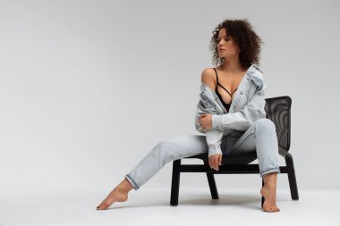 Kıvırcık saç stili, modaya uygun kot ceket ve kot pantolon giyen, beyaz stüdyoda sandalyede oturan, kendinden emin, genç, çıplak ayaklı bir manken.