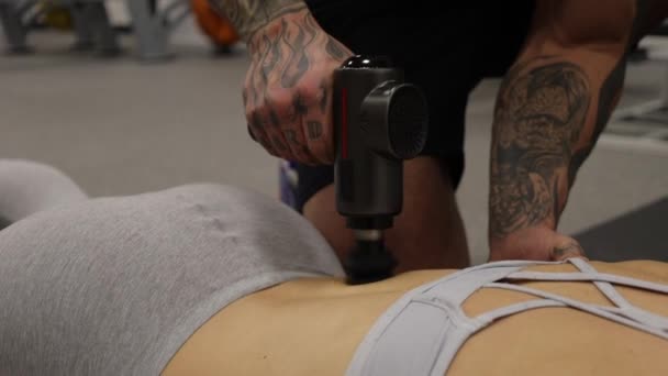 スポーツウーマンの体にマッサージ銃を使用したパーソナルトレーナー — ストック動画