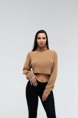 Bej ceketli, taytlı çekici bir bayan vücudu kucaklıyor ve beyaz arka planda kameraya bakıyor.