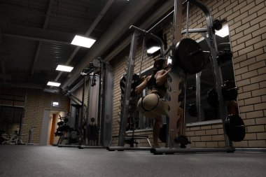 Spor kıyafetli, zayıf, seksi kalçalı, spor salonunda spor yaparken ağır halterli çömelme hareketleri yapan atletik bir kadının düşük açılı arka görüntüsü.