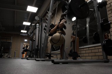 Spor kıyafetli, zayıf, seksi kalçalı, spor salonunda spor yaparken ağır halterli çömelme hareketleri yapan atletik bir kadının düşük açılı arka görüntüsü.