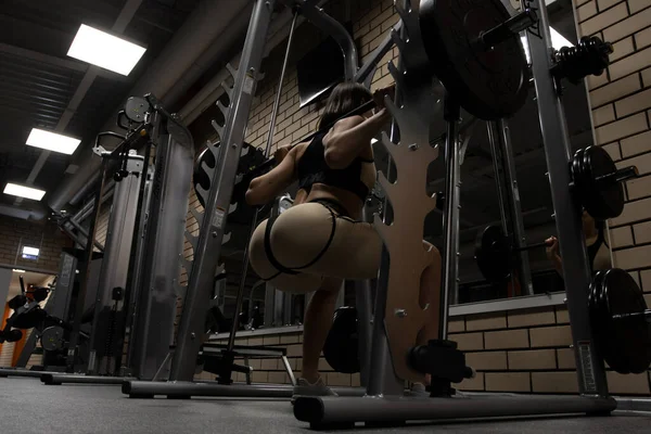 Spor kıyafetli, zayıf, seksi kalçalı, spor salonunda spor yaparken ağır halterli çömelme hareketleri yapan atletik bir kadının düşük açılı arka görüntüsü.