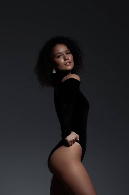 Siyah seksi iç çamaşırları ve yüksek topuklu ayakkabılar giyen uzun boylu kadın karanlık stüdyoda zarif bir şekilde kameraya bakıyor.