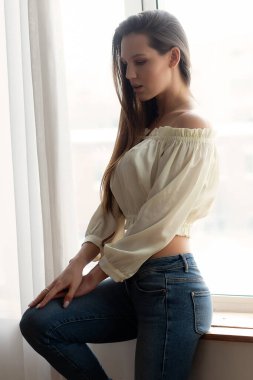 Şık bluzlu, kot pantolonlu, genç, sıska bir kadın. Kolunu kaldırıp, açık yatak odasının kapısına yaslanmış.