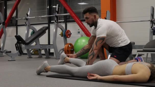 Personal trainer stretching legs of sportswoman in gym — Stock Video