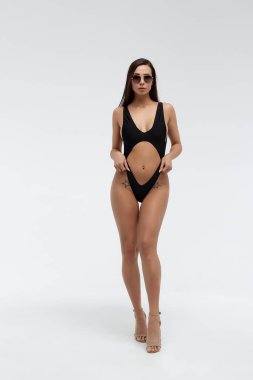 Çekici, uzun saçlı, esmer, seksi siyah bikinili ve modaya uygun güneş gözlüklü beyaz arka planda dururken saçlarına dokunan bir model.