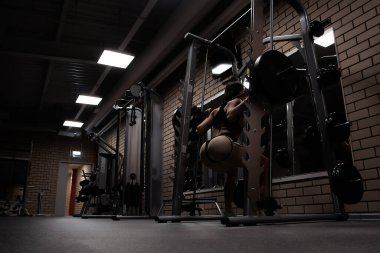 Spor kıyafetli, zayıf, seksi kalçalı, spor salonunda spor yaparken ağır halterli çömelme hareketleri yapan atletik bir kadının düşük açılı arka görüntüsü.