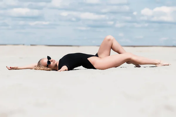 在风景如画的日子里 戴着太阳镜 身穿黑色泳衣的塑料女人优雅地坐在沙滩上伸展腿 — 图库照片