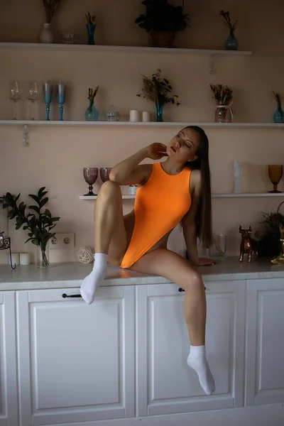 全长华丽的年轻黑发姑娘穿着性感的橙色紧身衣 坐在厨房里 俯瞰着四周 — 图库照片