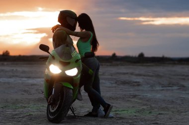 Birbirlerine aşık çiftin kumlu deniz kıyısında güneş batarken motosikletin yanında nazikçe kucaklaşmalarını yan görüyorum.